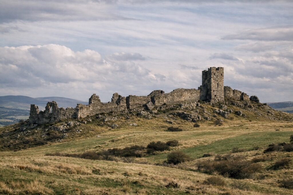Lange Burgruine mit Turm auf einem kahlen Rücken über Wiesen und Feldern, Hügel in der Ferne
