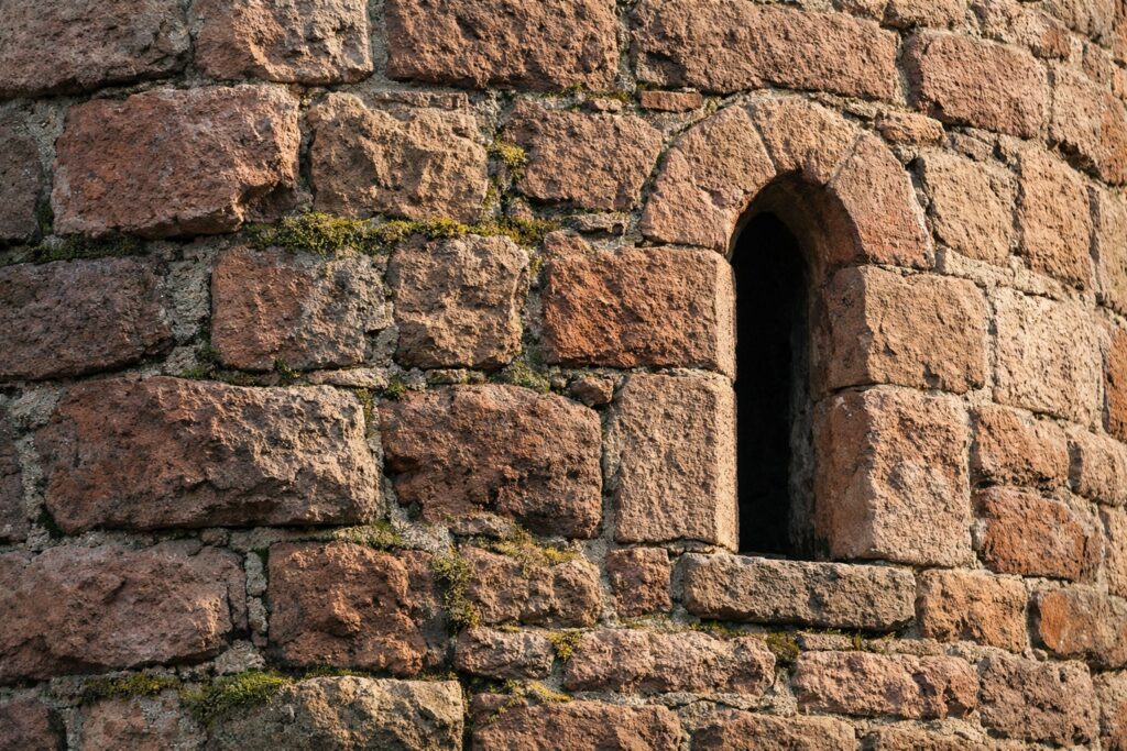 Nahaufnahme von verwittertem roten Sandsteinmauerwerk mit schmalem Rundbogenfenster, Moos in den Fugen